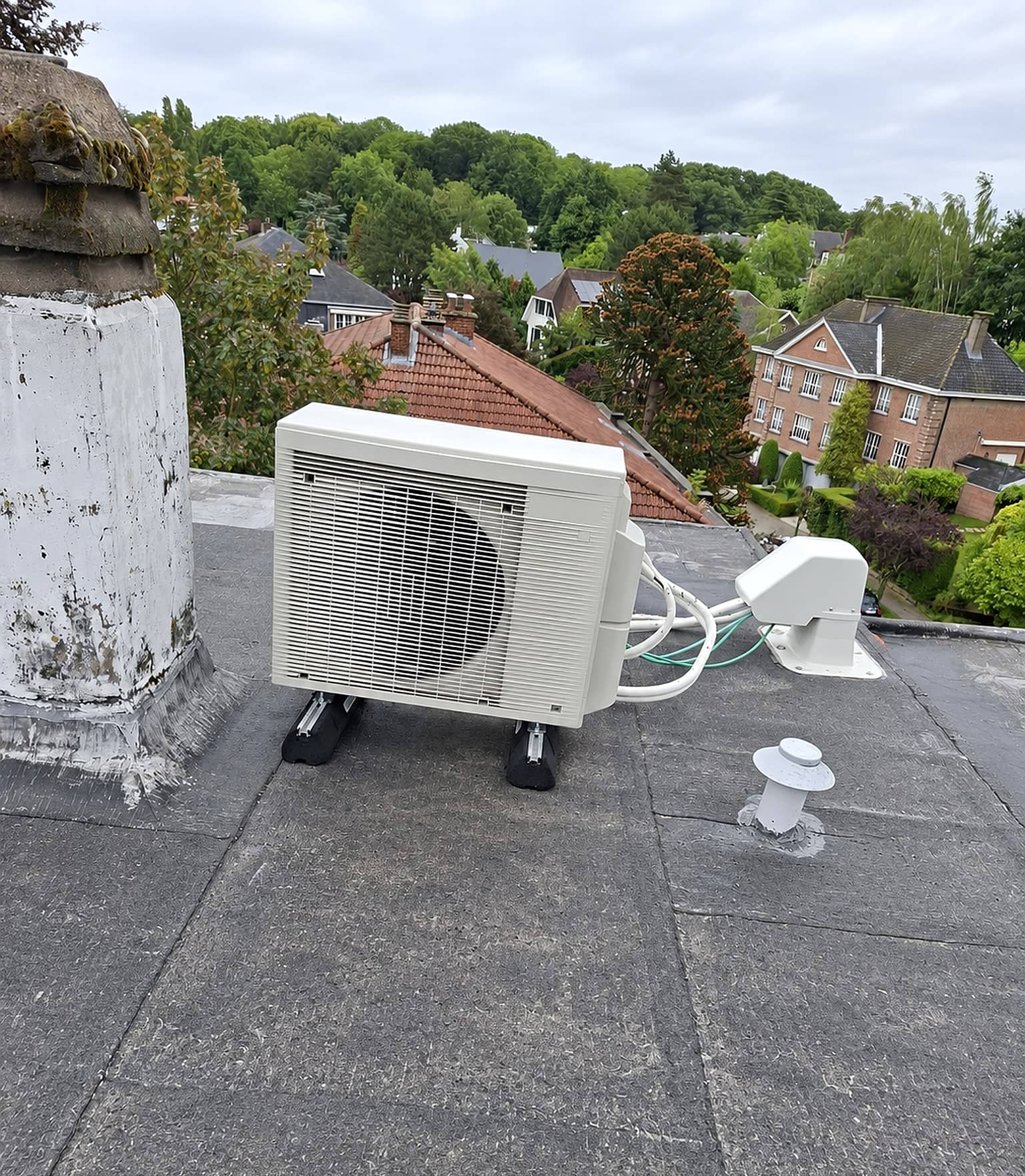 Groupe extérieur Daikin posé sur supports sur un toit plat, avec ses liaisons frigorifiques sous goulotte, maisons et arbres en arrière-plan